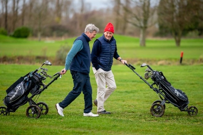Two older men smiling pulling golf buggies on a golf course